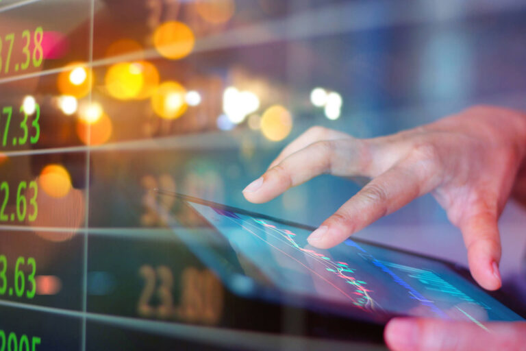 Image of hand working on tablet with financial charts and stock tickers in the background.