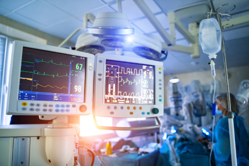 Photo showing two medical device monitors in the foreground and a medical caregiver checking additional medical devices in the background.